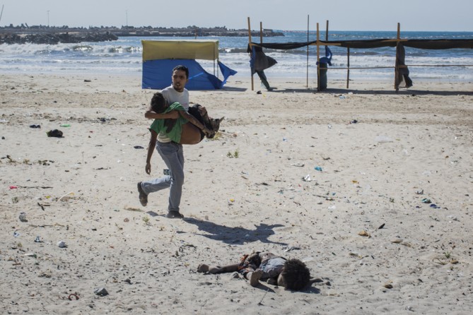 July 16, 2014. The body of a boy lies on a Gaza beach as a rescuer carries away another boy. Four boys died after two explosions, about 30 seconds apart. The first struck a small shack on a sea wall. The second, witnessed by the photographer, Tyler Hicks, from a nearby window, happened on the open beach near the boys as they were fleeing. Hicks rushed to the beach and photographed the scene. (Tyler Hicks/The New York Times)