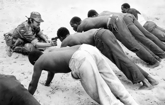 While interrogating prisoners at gunpoint, a white soldier makes suspected guerrillas hold a stress position for over an hour in the midday heat of the Kalahari desert on Rhodesia's western border with Botswana, 21 September 1977.