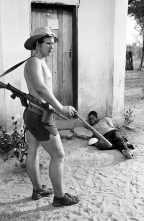 Lt. Graham Baillie raps a small wooden bat against his leg after using it to beat Moffat Ncube, a local teacher, political leader and now a bound, unconscious prisoner slumped against the wall of school house, 20 September 1977.  Ncube reportedly later died after three days of brutal, non-stop torture.