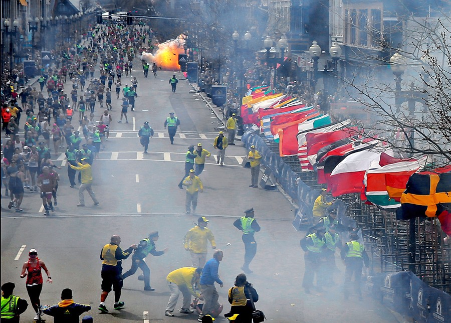 As police rush to the scene of the first blast near the finish line of the Boston Marathon, a second bomb explodes several blocks away. The shock wave from the first blast keeps the flags aloft lining the finish line. Behind the metal barricades, hundreds of people are injured and dozens fight for their lives. (Photo by David L. Ryan, April 19, 2013)